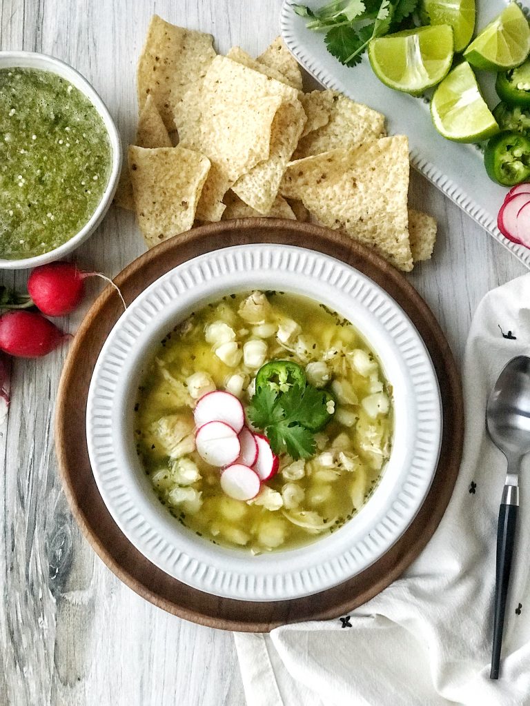 Bowl of pozole verde or green pozole. Radishes, cilantro, and jalapeno garnish. Salsa verde and tortilla chips on the side.