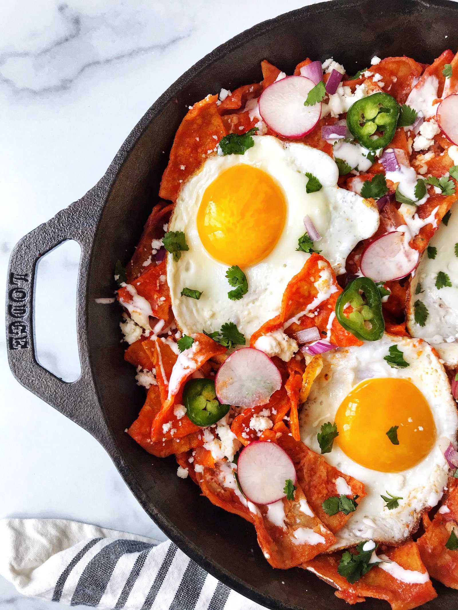 Chilaquiles Rojos (Red Chilaquiles) in cast iron skillet with cheese, sour cream, onions, jalapenos, fried eggs