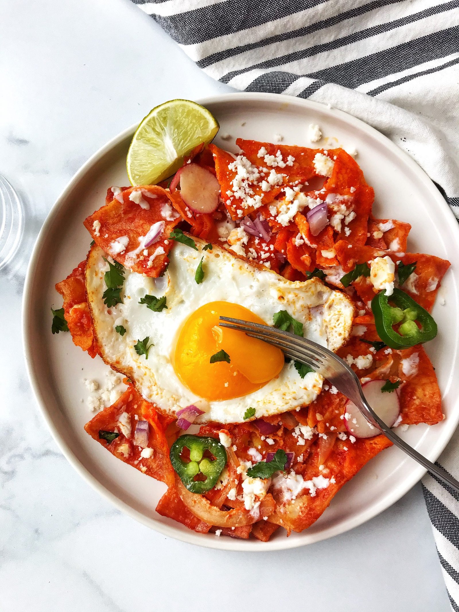 Chilaquiles Rojos (Red Chilaquiles) on white plate with cheese, sour cream, onions, jalapenos, fried egg with runny yolk