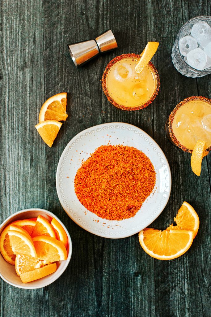 Orange mango margarita in glass with Tajin rim. Orange slices in bowl, Tajin on plate. Metal jigger and glass with ice shown.