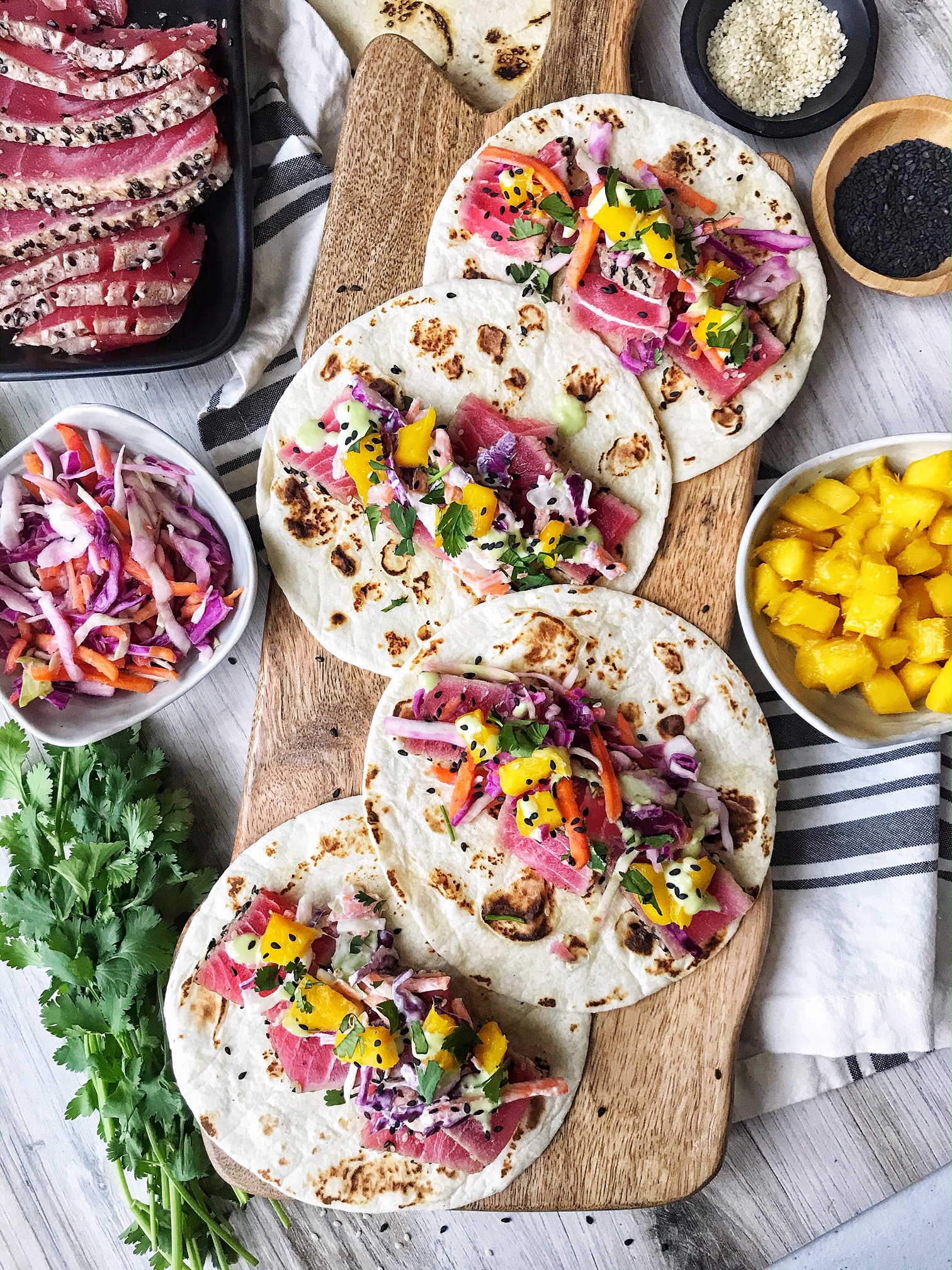 Ahi Tuna tacos with slaw and mango on top. Tacos on wood board and slaw, cilantro, sesame seeds, and mango on the side