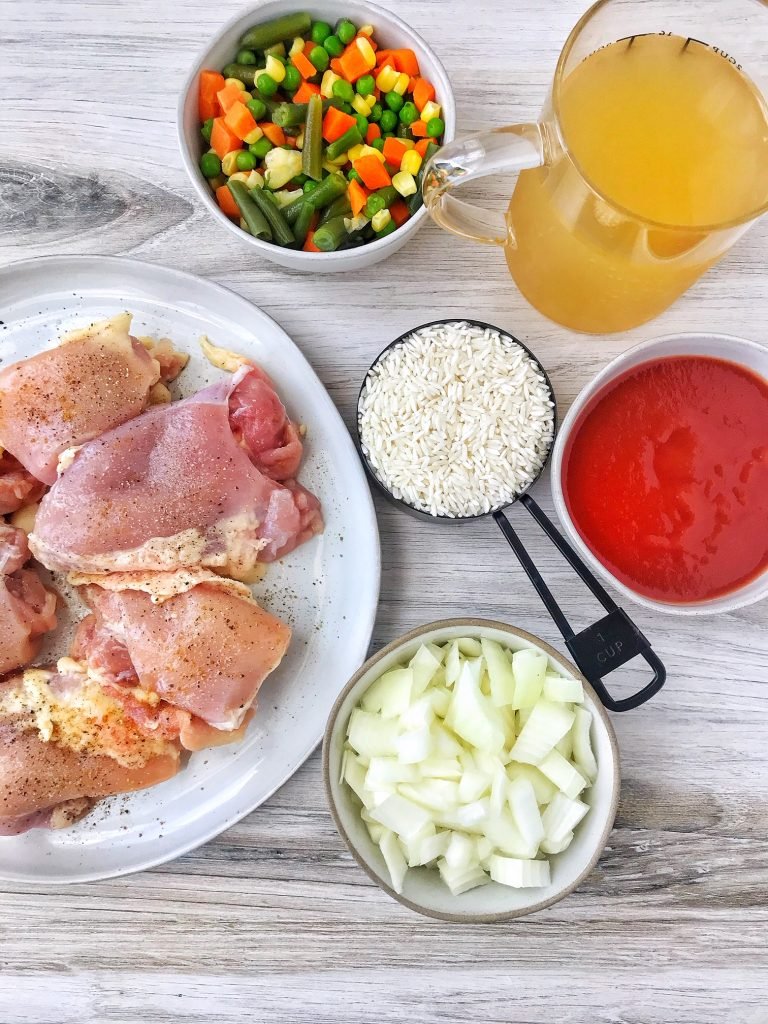 Ingredients for arroz con pollo. Plate of chicken thighs, bowl of mixed vegetables, cup of rice, cups of chicken broth, bowl of tomato sauce, bowk of chopped onion.