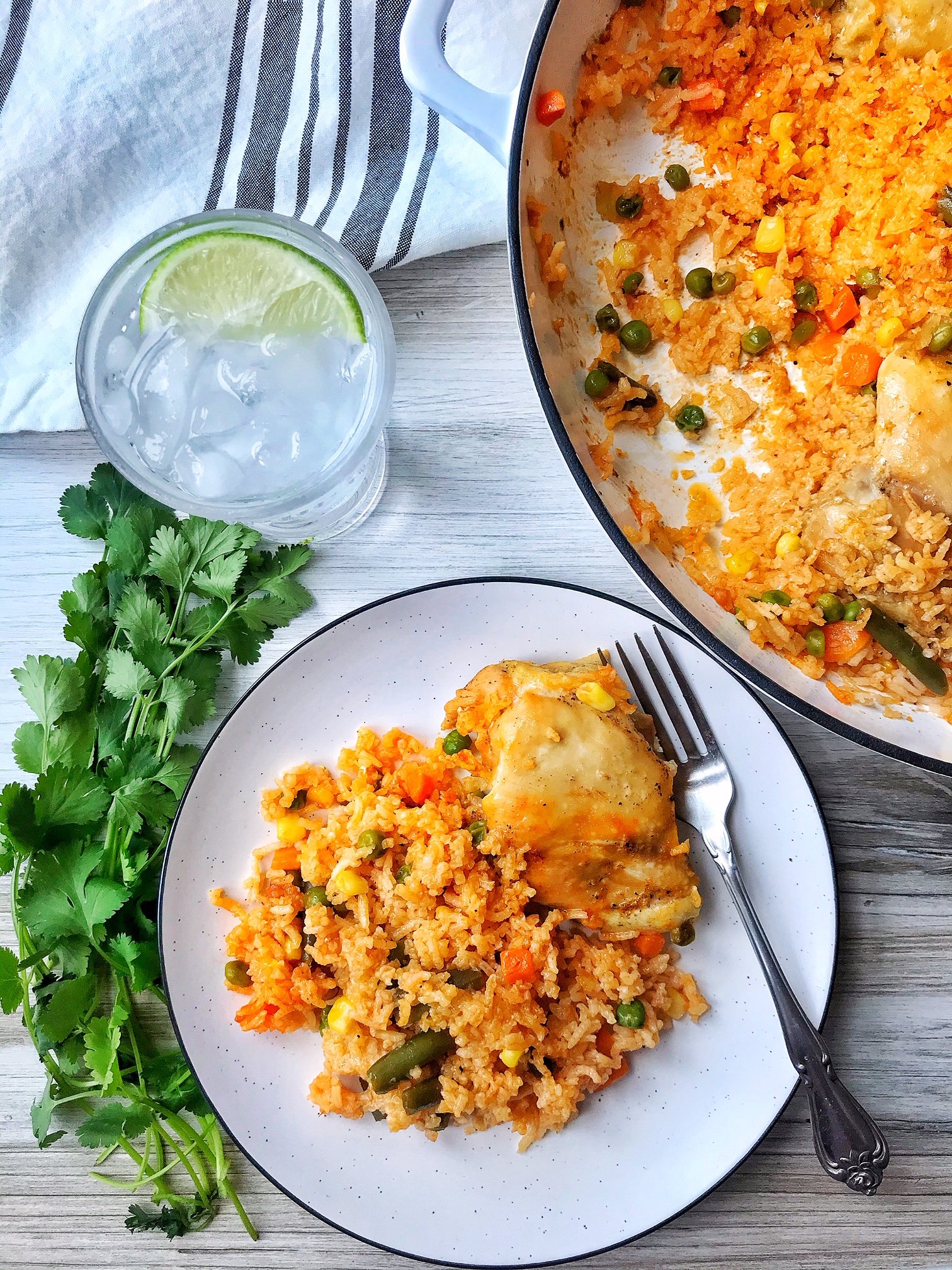 Large pan of chicken and rice. Small plate with chicken thigh and rice with vegetables. Glass of iced water with lime.  Cilantro on the side.