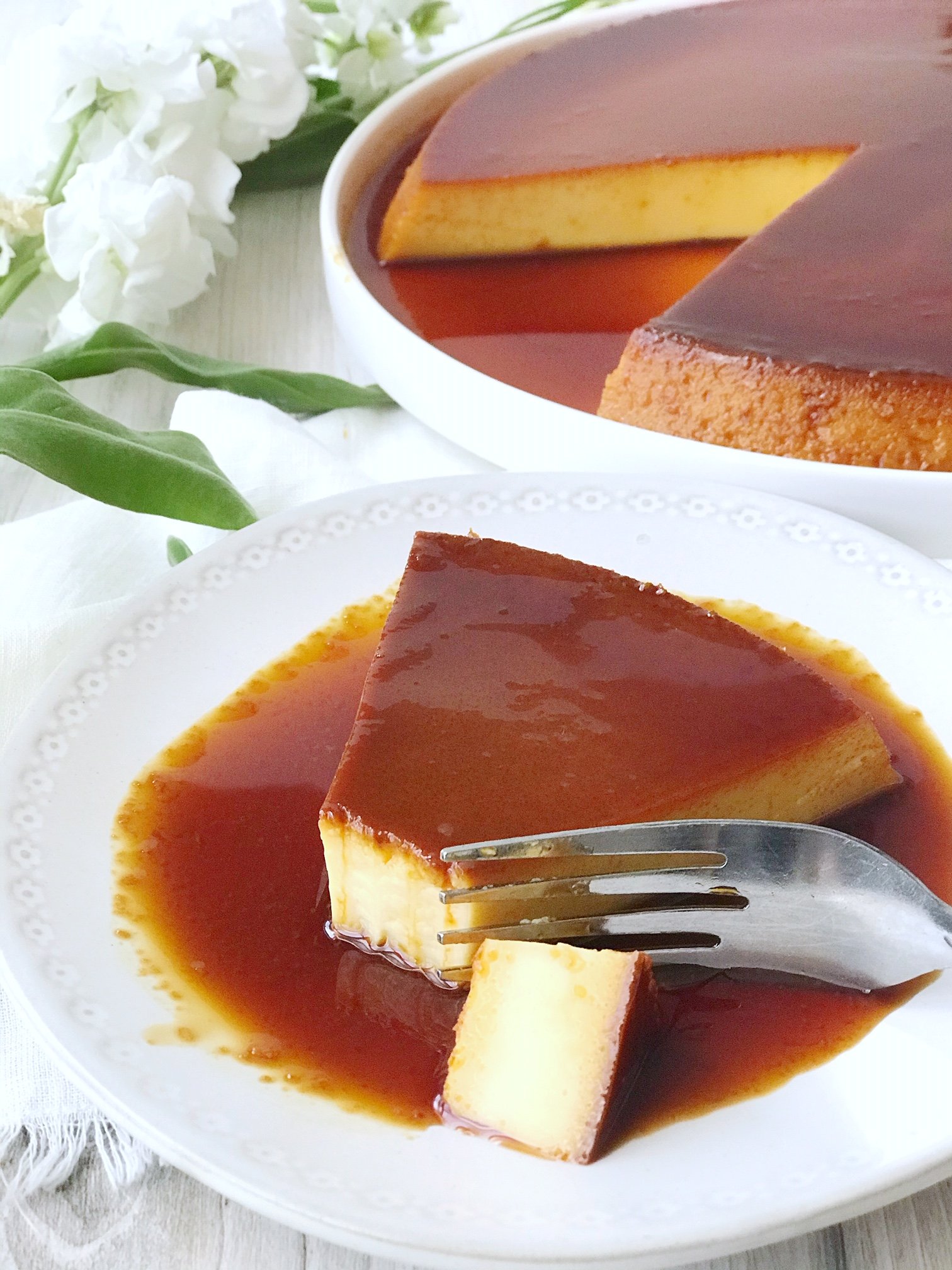 Mexican Flan on a large plate with caramel sauce and a slice missing. Slice of Mexican flan on small plate with piece cut off and a fork. White flowers in background.