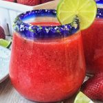 Frozen strawberry margarita in glass with salted rim garnished with lime. Strawberries in background
