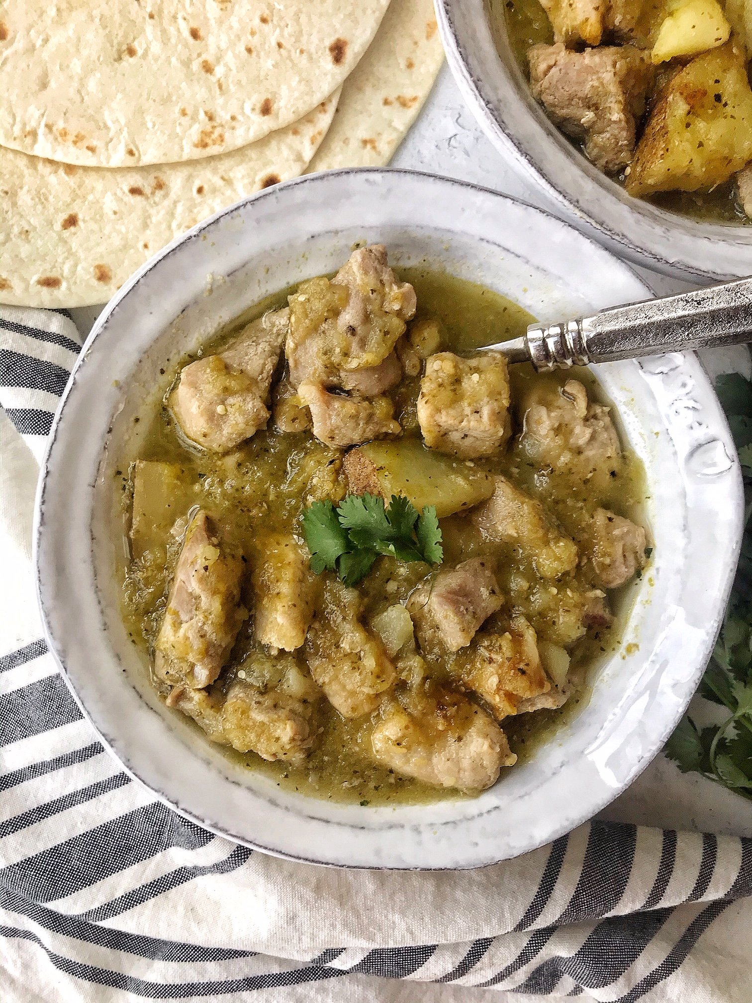 White bowl with pork chile verde and silver spoon.  Flour tortillas on the side. 