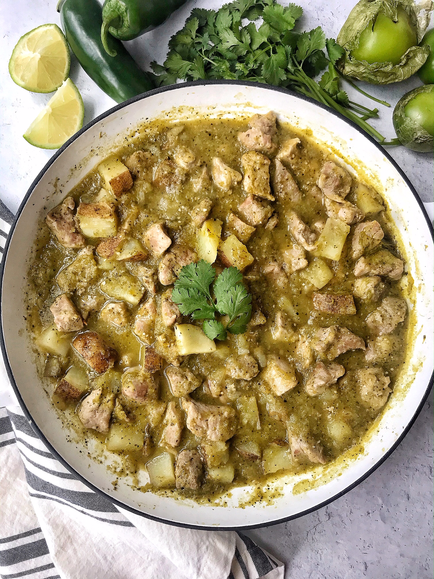 Pork chile verde in a white skillet.  Jalapenos, tomatillos, lime, and cilantro as props