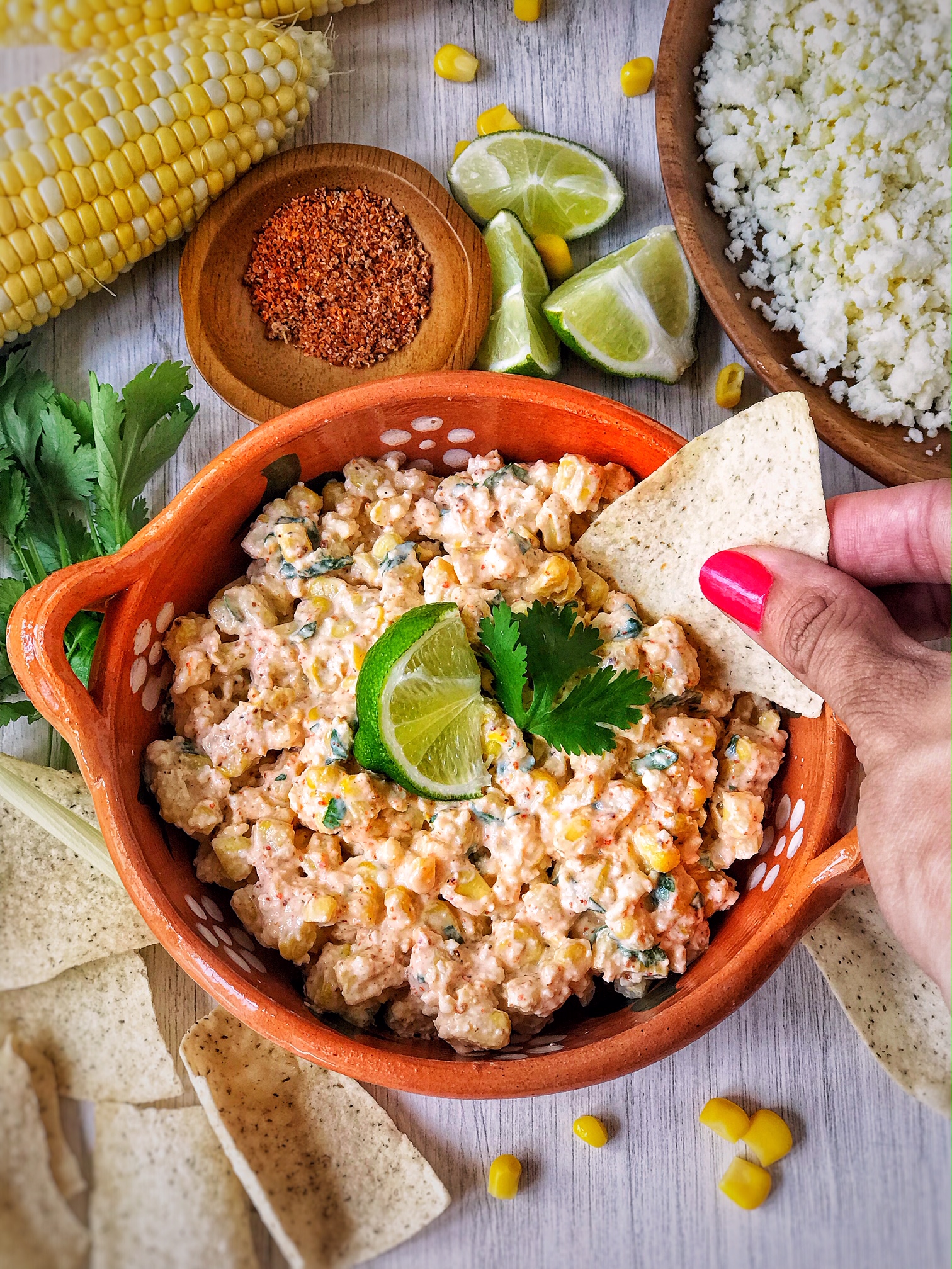 Bowl of Mexican street corn dip garnished with lime and cilantro. Hand in picture with tortilla chip in dip. Corn, limes, and crumbled cheese in picture.