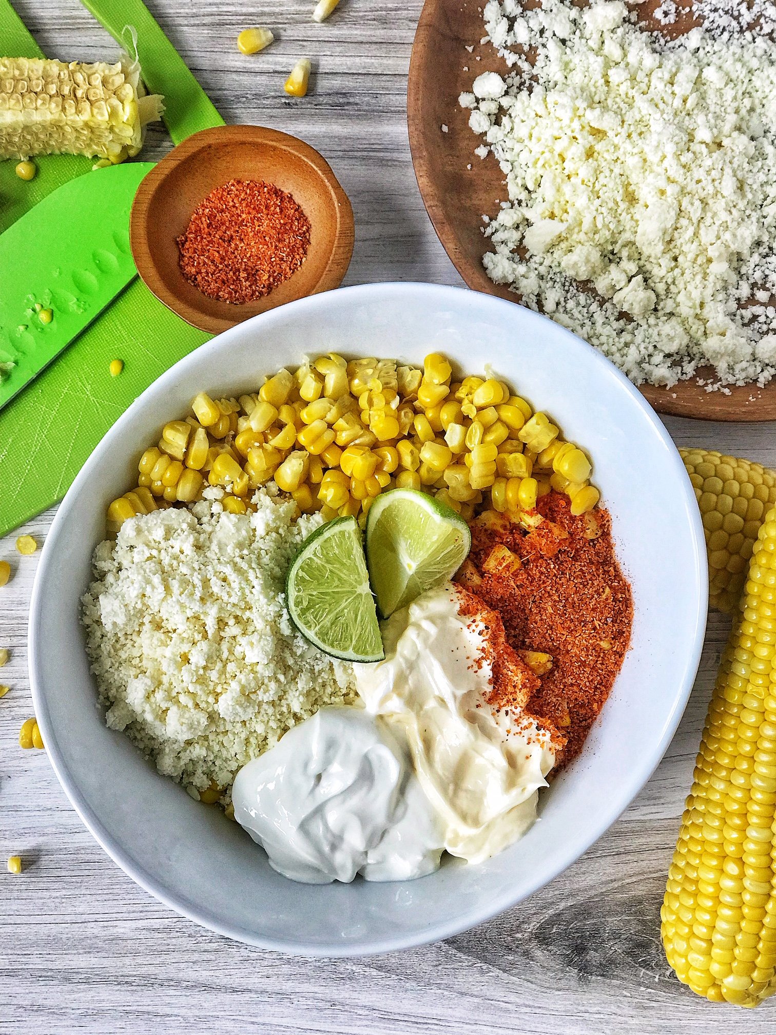 White bowl with Mexican street corn dip ingredients. White bowl with corn, crumbled cheese, mayo, sour cream, Tajin, and lime. Corn and crumbled cheese in picture.