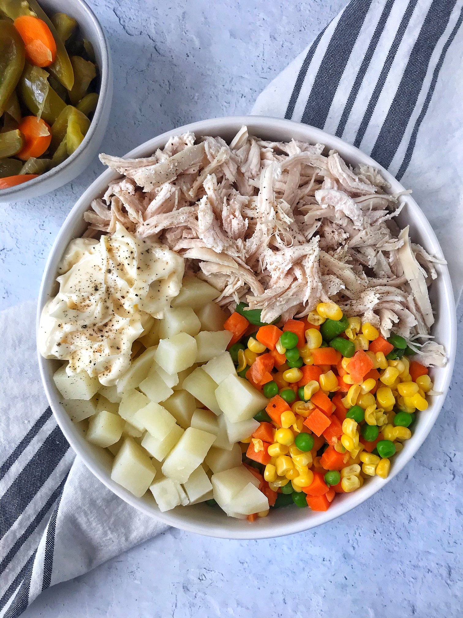 White bowl with shredded chicken, mayo, pepper, potatoes, and mixed vegetables. Small bowl with pickled jalapeños on the side.