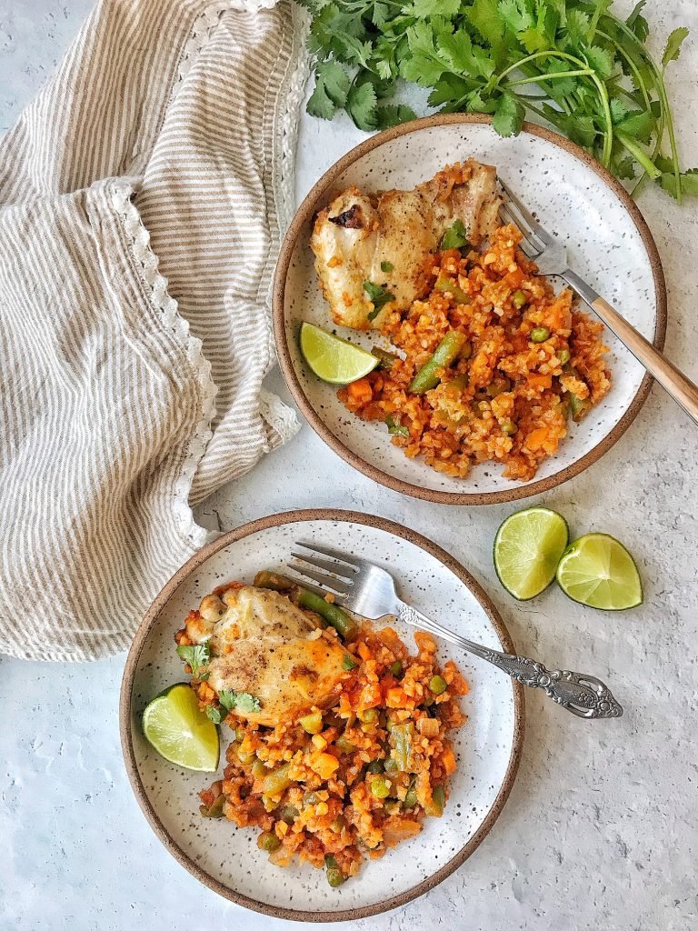 Two white plates with chicken thigh, cauliflower rice with vegetables in a tomato sauce, and lime wedges on the side. Garnishes with cilantro.