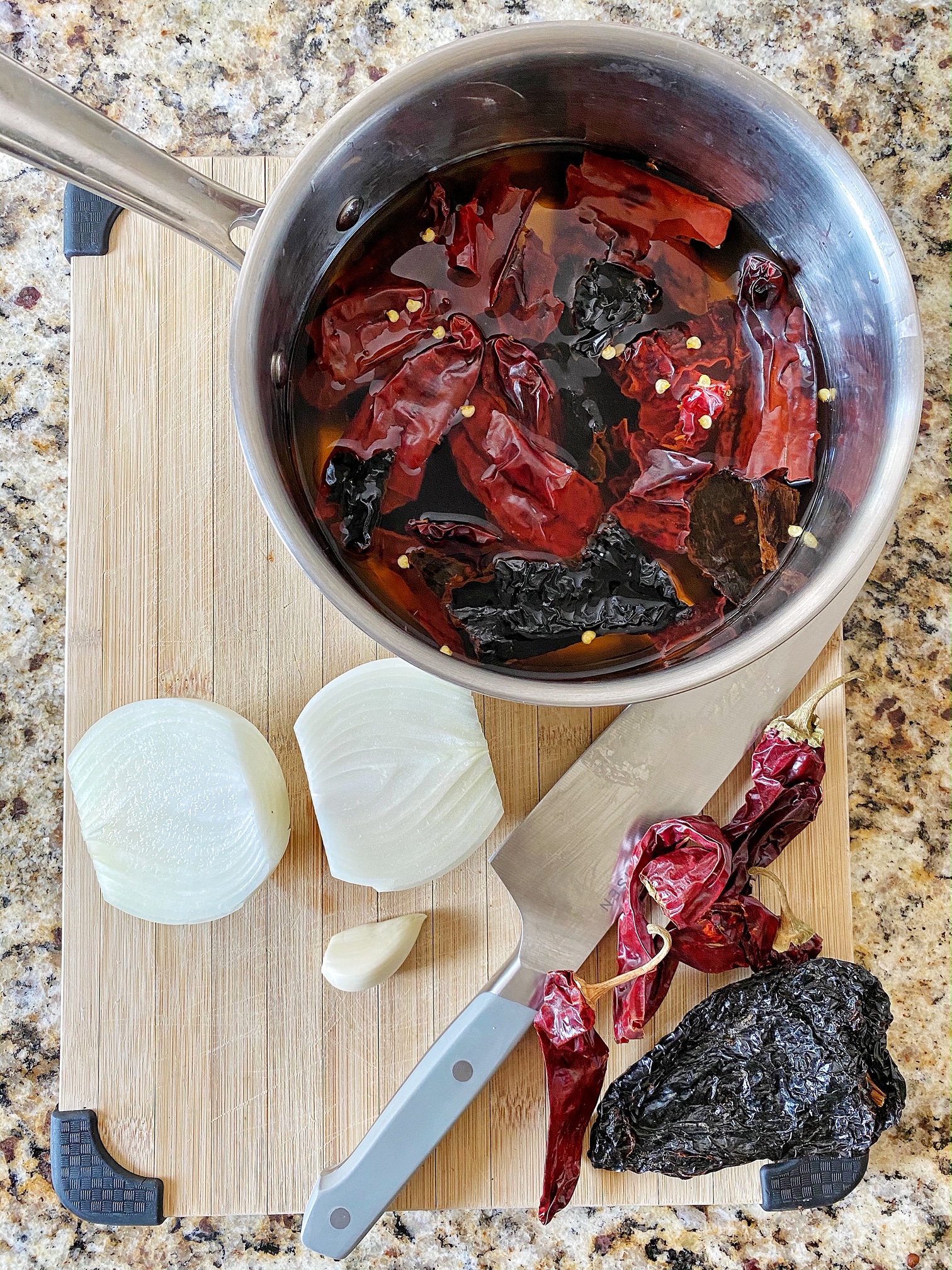 Saucepan with water and dried peppers. Onion, garlic, and dried peppers on cutting board.