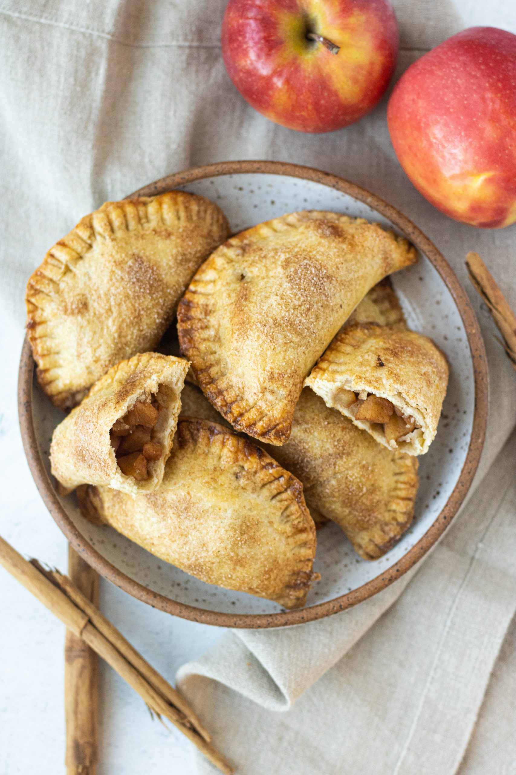 White bowl with brown trim full of cinnamon apple empanadas. Apples and cinnamon sticks as garnish. 