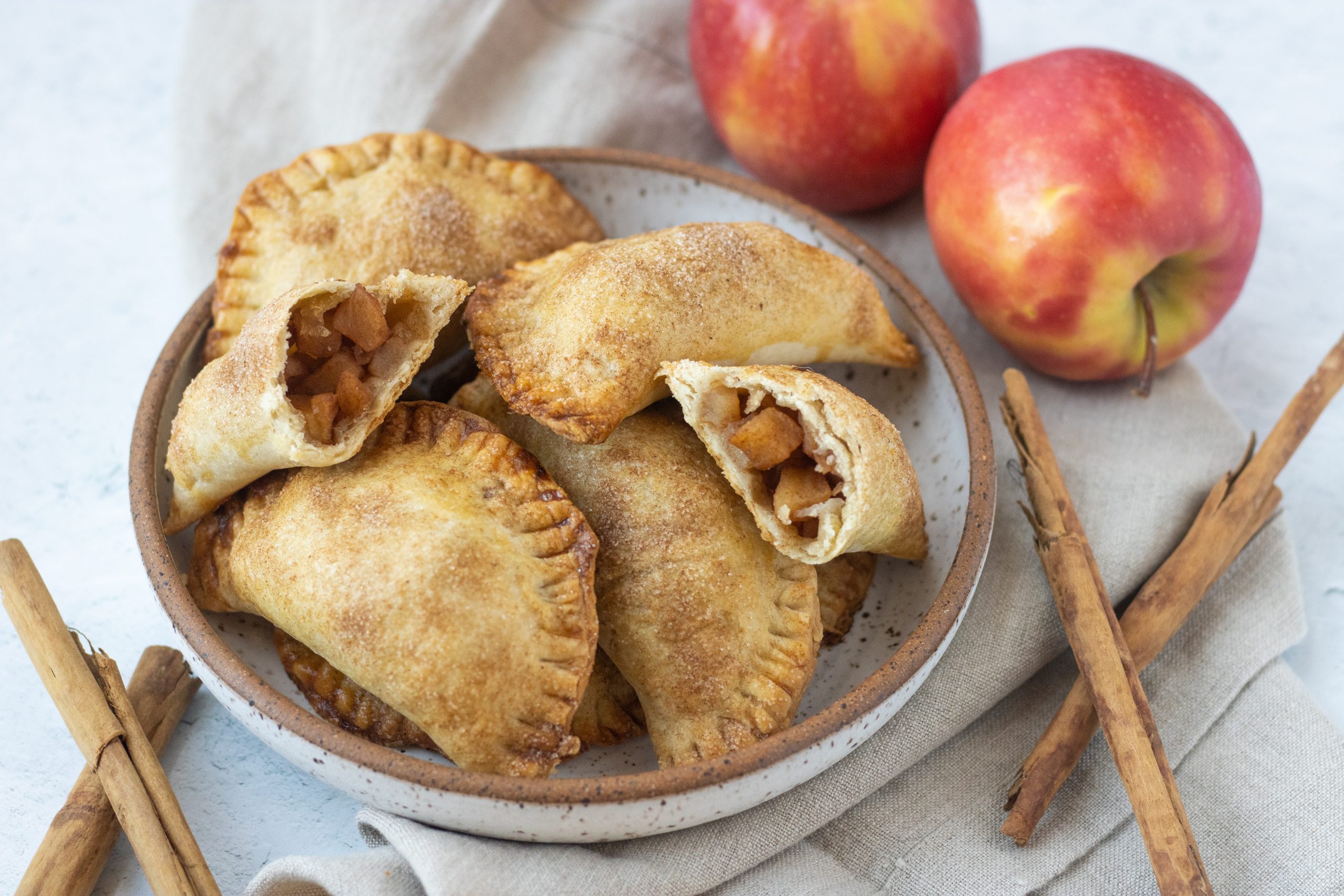 White bowl with brown trim full of cinnamon apple empanadas. Apples and cinnamon sticks as garnish. 