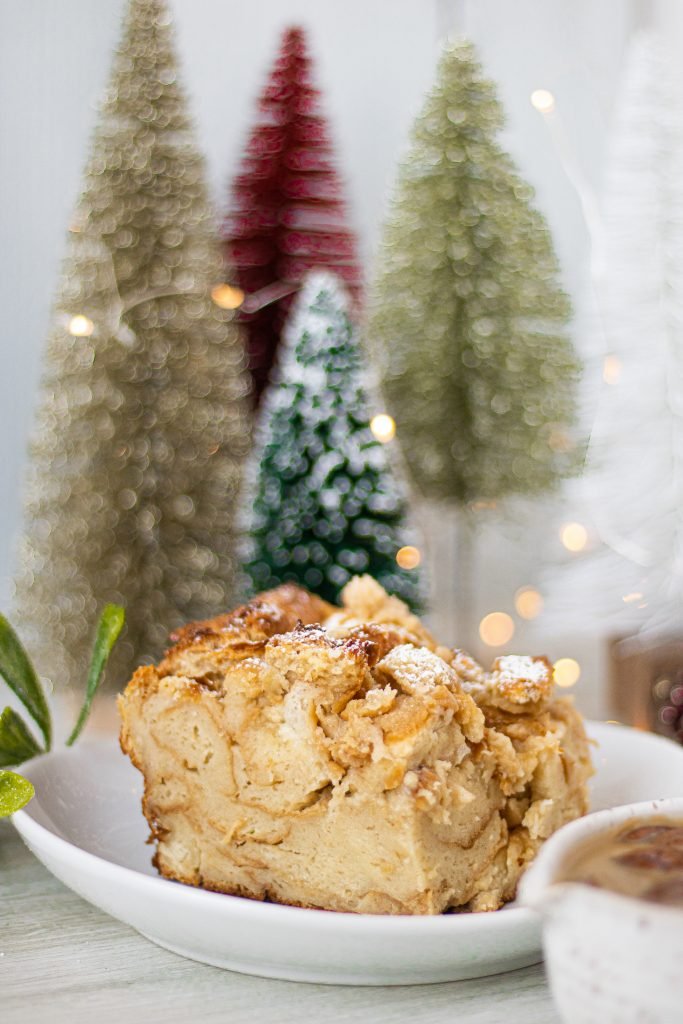 Slice of bread pudding in white bowl sprinkled with powdered sugar. Background with Christmas trees