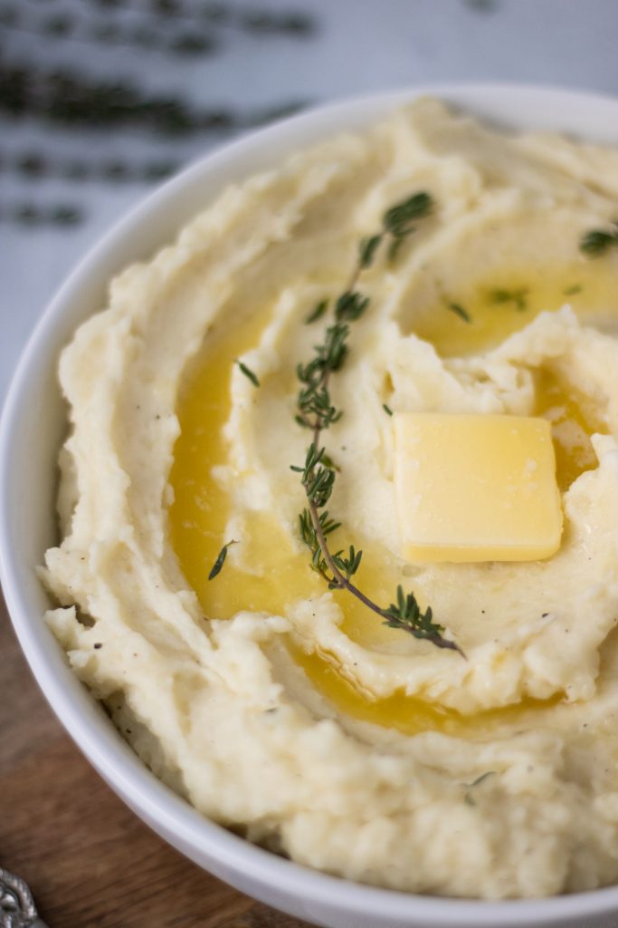 White bowl of creamiest mashed potatoes with butter and thyme sprigs