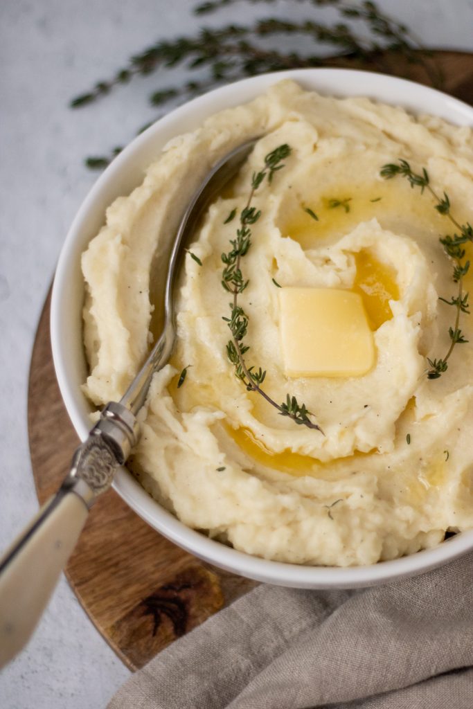 white bowl of mashed potatoes with butter and thyme sprigs. Serving spoon in mashed potatoes.