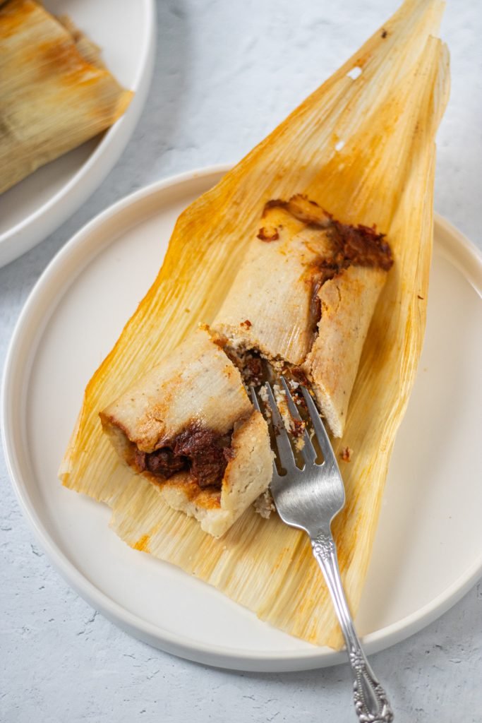 One authentic mexican tamale on corn husk on top of white plate. Fork on side.