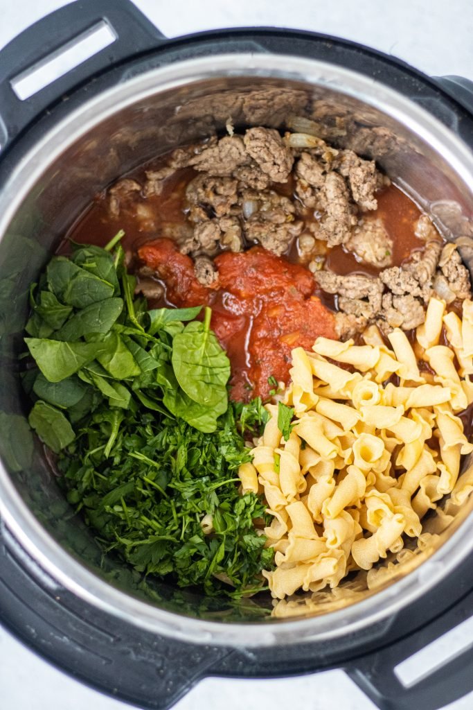 Instant pot with lasagna soup ingredients. Pasta, Spinach, parsley, pasta sauce, sausage.