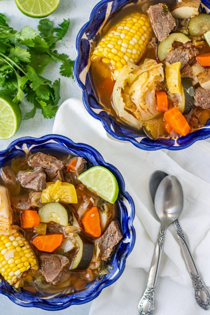 Blue bowls with Caldo de Res with zucchini, squash, corn, carrots, and beef. Cilantro and lime garnish