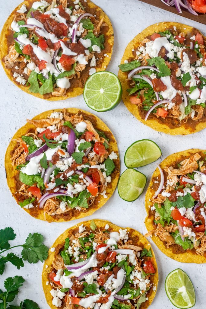 Multiple chicken tinga tostadas all topped with lettuce, onion, tomato, sour cream, cheese, and salsa. Limes and cilantro garnish.