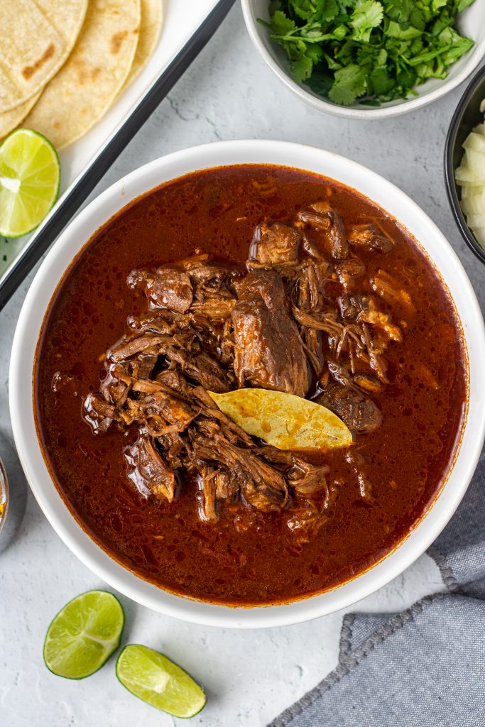 White bowl with birria de res (beef birria) with bay leaf. Lime garnish, bowl of cilantro, bowl of onion, and tortillas in background.