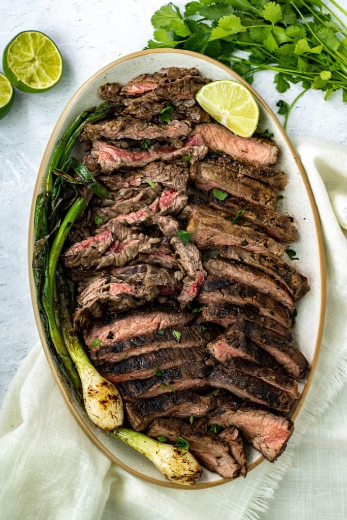 Slice Mexican Carne asada on platter with green onions, lime, and cilantro garnish,