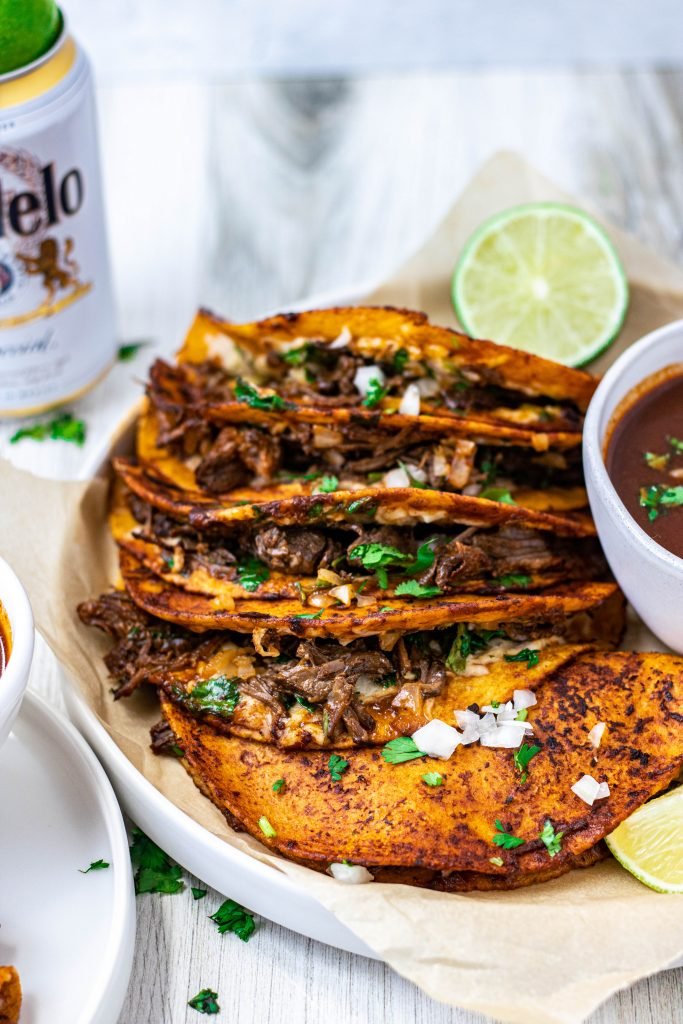Plate of quesabirria tacos with onion and cilantro on top, lime garnish, and beer on side