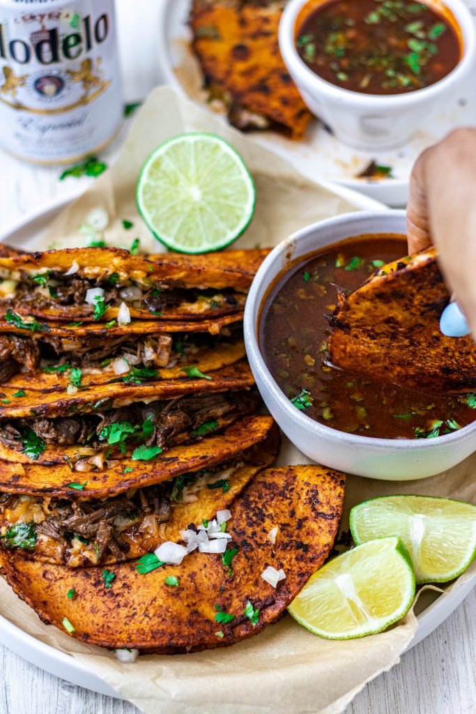 Plate of Quesabirria Tacos (Birria Quesadillas) with onion, cilantro, and lime garnish. Side of dipping sauce, or consomé, with quesabirria being dunked into it. Beer on side