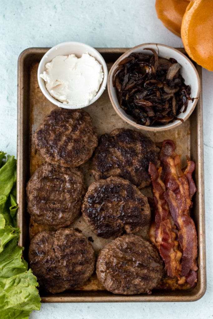 Ingredients for burgers including goat cheese, caramelized onions, burger patties, bacon, buns, and lettuce