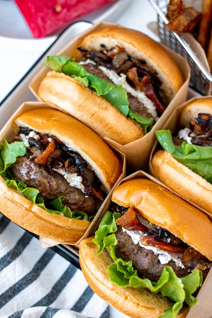 Close up of four Bacon Goat Cheese Burgers with Balsamic Caramelized Onions. Burgers wrapped in brown paper.
