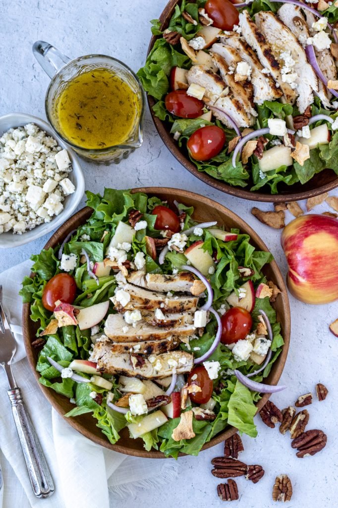 Chicken and Apple Salad in a wooden bowls with apples, tomatoes, red onion, grilled chicken, gorgonzola cheese. Small bowl of gorgonzola cheese, dressing on side. Apple and pecans for garnish